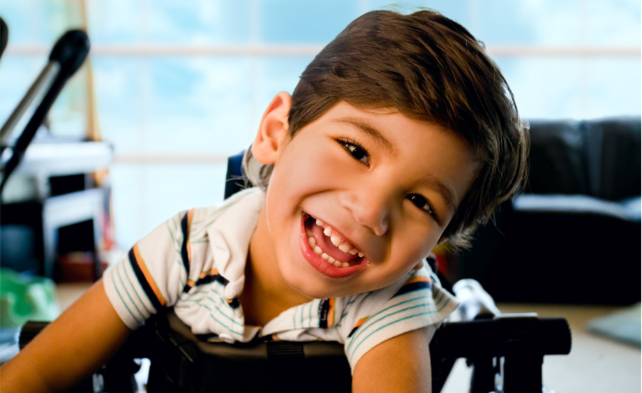 Disabled little boy standing in walker smiling and happily looking at camera with bright blue sky and clouds in the background window