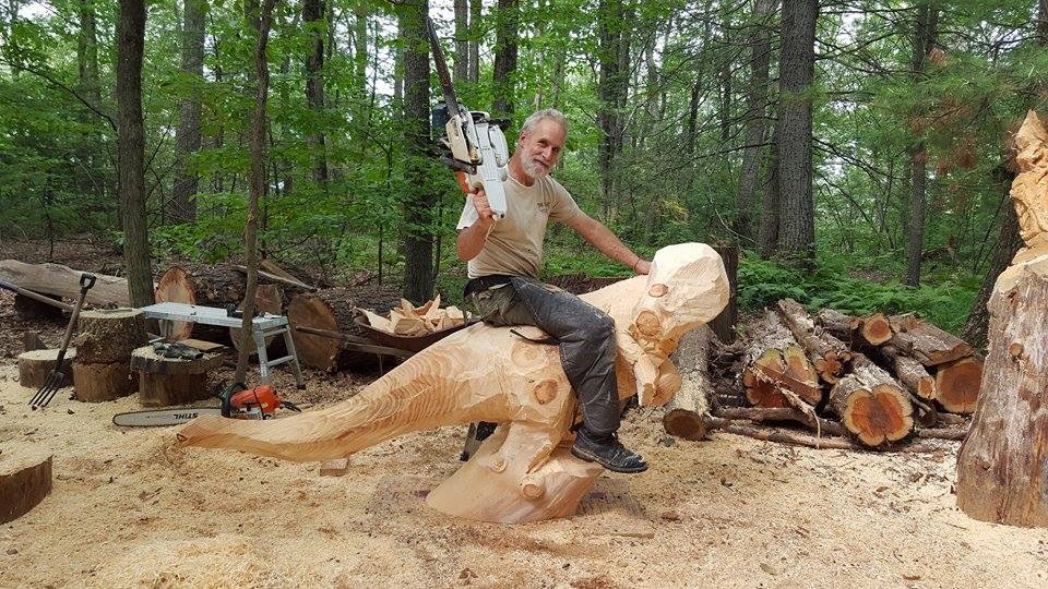 Todd Gladfelter holding a chainsaw and sitting on a dinosaur he carved out of wood