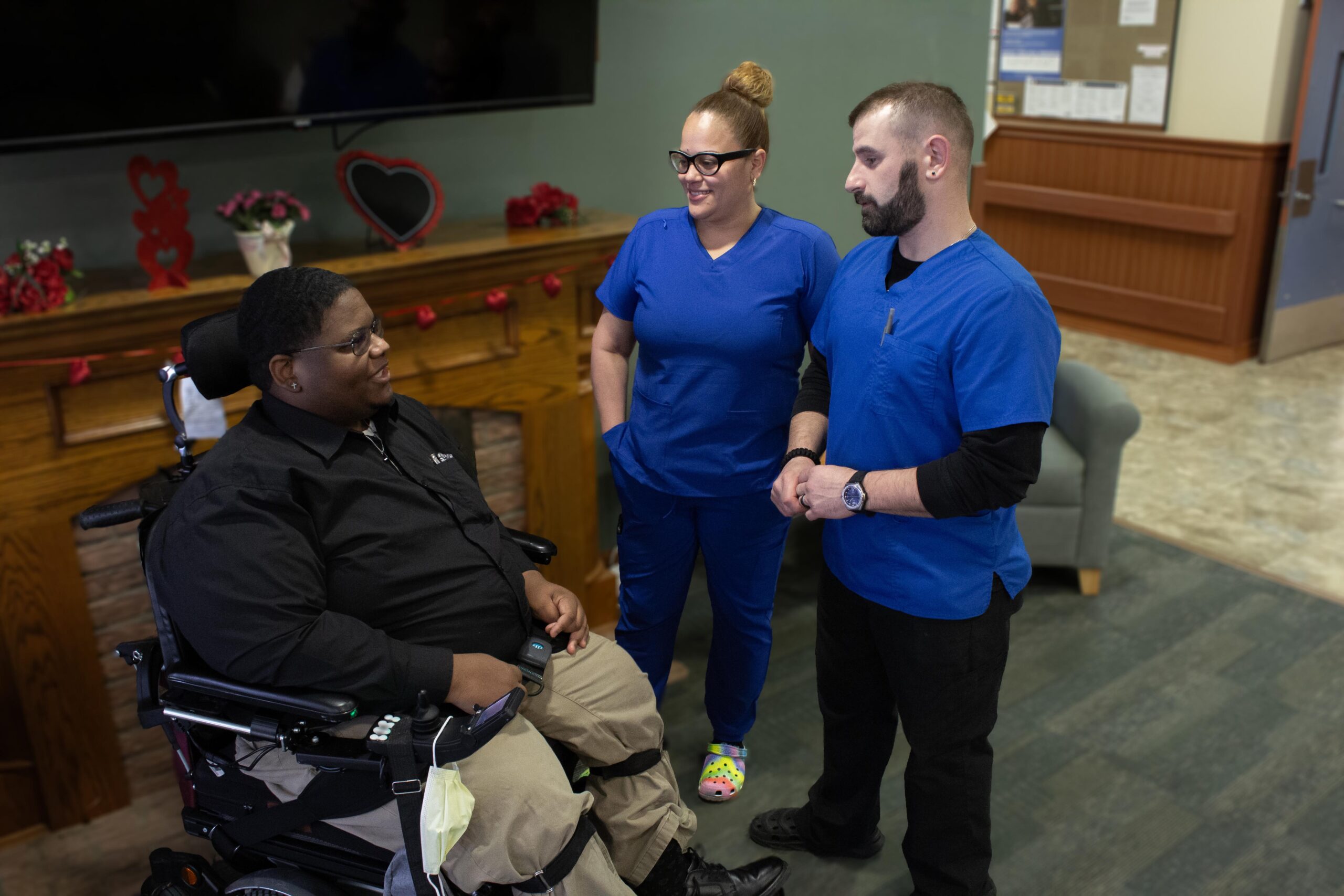 Two nurses in blue scrubs are talking to a person in an a person that is in a wheelchair.